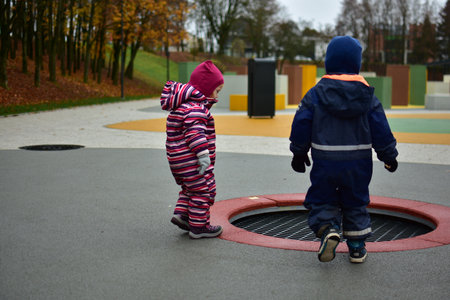 Two toddlers dressed in warm winter suits standing near a sunken playground trampoline on a colorful outdoor play area, enjoying autumn weather together. Siblings having fun outside.の写真素材