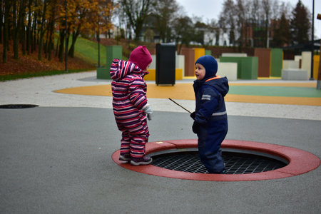Two toddlers dressed in warm winter suits standing near a sunken playground trampoline on a colorful outdoor play area, enjoying autumn weather together. Siblings having fun outside.の写真素材