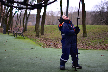 Toddler boy looking up and holding rope playground element in autumn park, wearing warm winter...