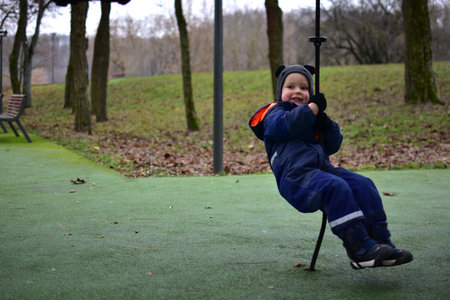 Toddler boy hanging and playing on rope playground in autumn park. Child wearing warm winter clothes...