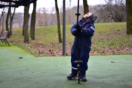 Toddler boy looking up and holding rope playground element in autumn park, wearing warm winter clothes. Outdoor play supports balance, coordination and motor skills development.の写真素材