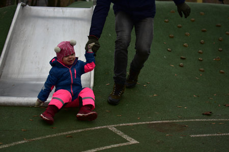 Happy toddler girl sliding down playground slide while holding adult hand. Child enjoys safe outdoor play with parent support in autumn park.の写真素材