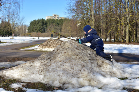Toddler climbing a dirty snow pile in a sunny winter park while playing with a stick. Curious child...