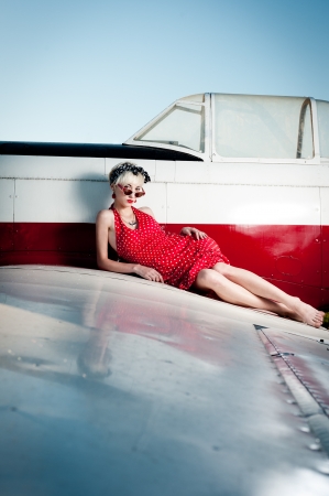 outdoors pin-up portrait of young blond sexy caucasian woman in red retro dress lying on airplane wingの写真素材
