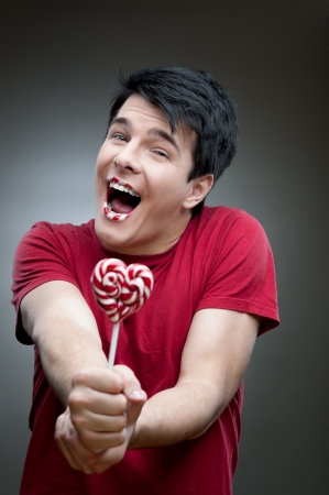 young funny caucasian brunette man in red t-shirt holding lollipop over gray backgroundの写真素材