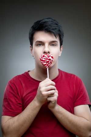 young funny caucasian brunette man in red t-shirt holding lollipop over gray backgroundの写真素材