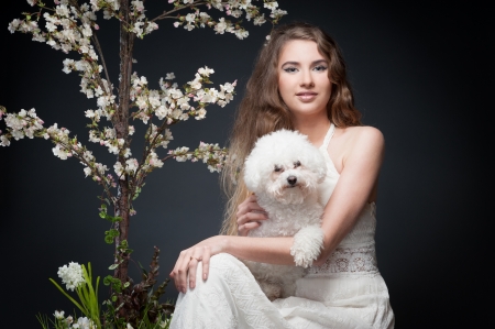 sensual caucasian woman in white dress and puppy sitting near flowering tree over gray backgroundの写真素材