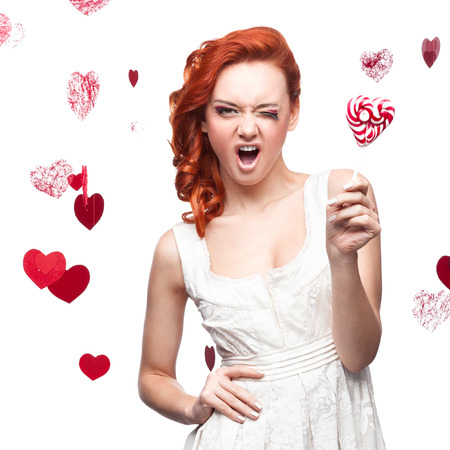 cheerful caucasian red-haired winking young woman holding lollipop. isolated on whiteの写真素材