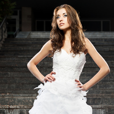 outdoors portrait of beautiful young caucasian brunette woman in white wedding dress over gray stairs on backgroundの写真素材