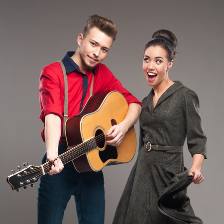cheerful young caucasian guitar player and dancing girl in vintage clothing over gray background