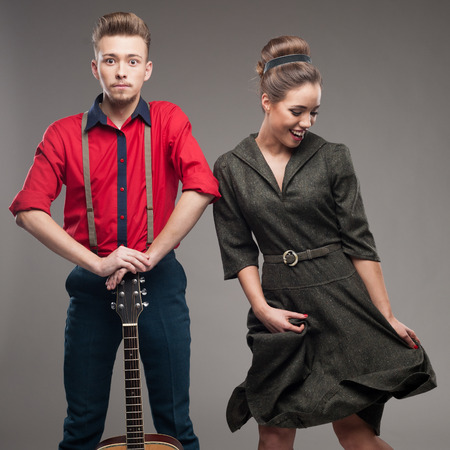 cheerful young caucasian guitar player and dancing girl in vintage clothing over gray background