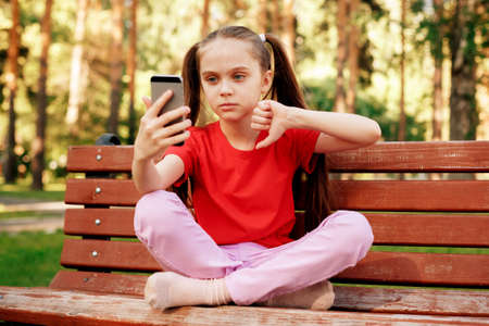 Little Girl with Smartphone Taking Selfie or Having Video Call And Showing Thumb Down In Park. Children and Technology Concept.の写真素材
