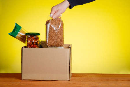 Volunteer with donation box with foodstuffs on yellow background