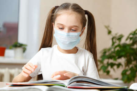 A little girl in a medical mask reading a book. Quarantine, stay at home. The who's recommendation. Coronavirus Pandemic Conceptの写真素材