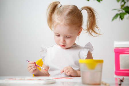 A Little Girl, With Tails On Her Head, Draws Watercolors Sitting At a Table. The Child Likes To Be Creative.の写真素材