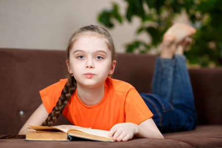 Cute caucasian little girl with a long braid reading book while lying on sofa at home during quarantine due to coronavirus. Education and leisure concept.の写真素材