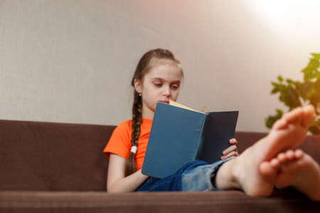 A funny little caucasian girl in an orange t-shirt with bare feet Reads an interesting book while sitting on a sofa at home. Foot closeup. selective focusの写真素材
