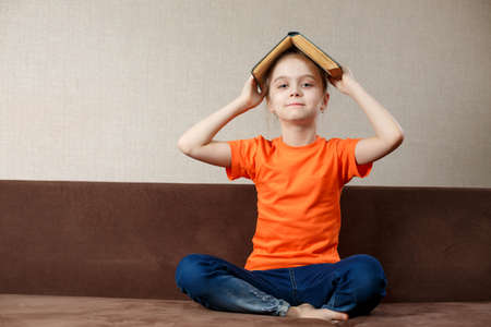 Happy little schoolgirl with book on her head, wearing orange t-shirt and jeans, sitting in sofa at homeの写真素材