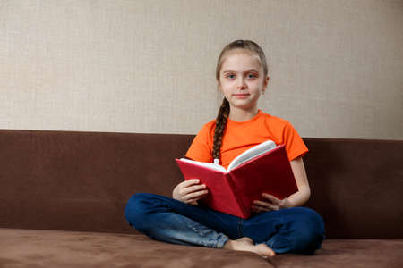 Caucasian Little Girl Sitting On The Sofa Reading a Book At Home.の写真素材