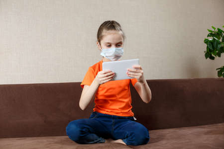 Quarantine Home Leisure.Little Girl In Medical Mask Using Tablet Computer Watching Movie Online Sitting on Sofa Indoorsの写真素材