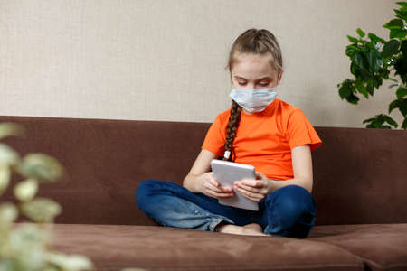 Little girl in medical mask sitting on the sofa in the Lotus position and using tablet pc. Isolation at home. Looking into the camera.の写真素材