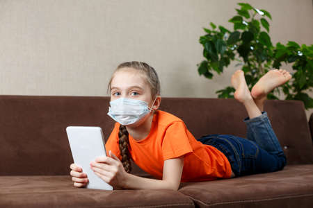 Little girl in medical mask lying on the sofa and using tablet pc. Isolation at home. Looking into the cameraの写真素材