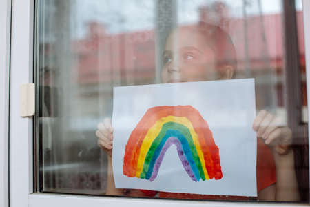 Little girl in the window holds a picture of a rainbow symbolizing the imminent end of the quarantine in connection with the coronavirus pandemicの写真素材