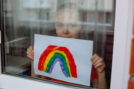 Adorable little toddler girl looks through the window with drawn rainbow in hands symbolizing the imminent end of the quarantine in connection with the coronavirus pandemicの写真素材