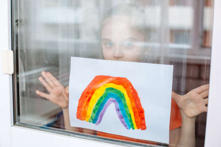 Little girl looking at infinity through a window and showing a picture of a rainbow during coronavirus quarantineの写真素材