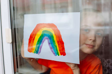 Little girl in the window holds a picture of a rainbow symbolizing the imminent end of the quarantine in connection with the coronavirus pandemicの写真素材
