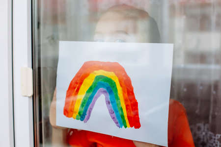Adorable little toddler girl looks through the window with drawn rainbow in hands symbolizing the imminent end of the quarantine in connection with the coronavirus pandemicの写真素材