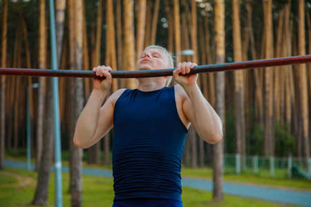 Young muscular build man doing pull ups exercises on horizontal bar outdoors, strong athletic runner...