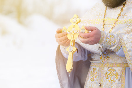 The hands of a priest dip an Orthodox gold cross into the river. Feast of the Epiphany of Russia