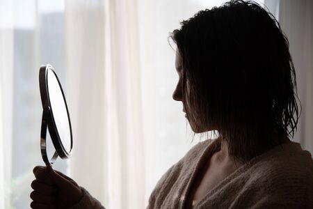 caucasian woman looking in mini mirror light window background.の写真素材