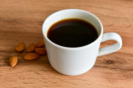 White cup of strong black coffee with almonds lying nearby on a wooden surface close-up.の写真素材