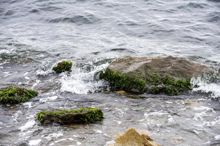 Coast with large stones in the sea water. A white wave of foam rolls onto the shore, washing several large black stones.の写真素材