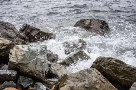 Coast with large stones in the sea water. A white wave of foam rolls onto the shore, washing several large black stones.の写真素材