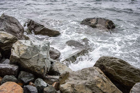 Coast with large stones in the sea water. A white wave of foam rolls onto the shore, washing several large black stones.の写真素材