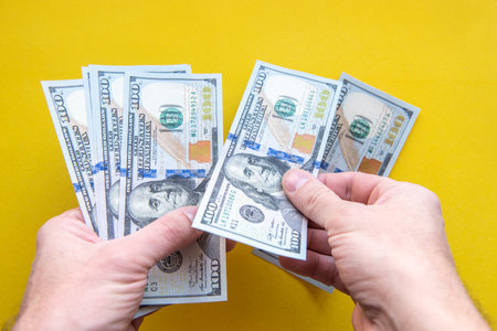 Male hands holding and counting one hundred dollar bills close-up on a yellow background.の写真素材