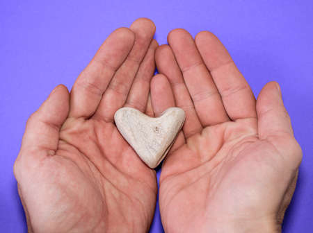 Male hands hold a heart-shaped stone on a purple background, close-up. romantic valentine's day. stone heart as a gift.の写真素材