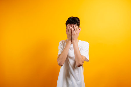 Teenager covering face with hands, yellow background.の写真素材
