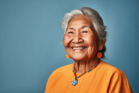 Smiling elder Inuit woman in casual wear, posing against a blue studio backgroundの素材