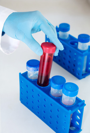 Hand in a blue rubber glove holding red laboratory container with a liquidの写真素材