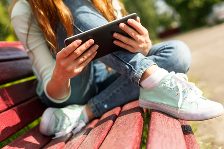 Younf woman with a black tablet sitting on a bench in a park. Close up shot of hands holding black tabletの写真素材