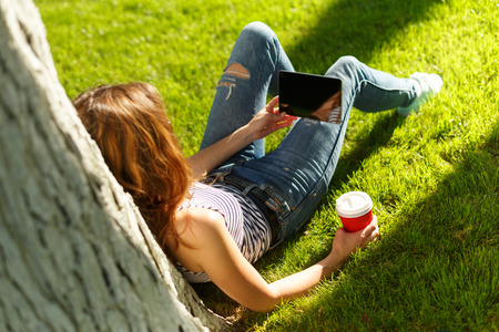 Young woman with red disposable paper cup of coffee using tablet pc under the tree on a green lawn in a parkの写真素材
