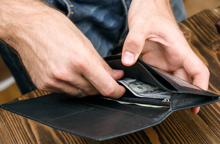 man is putting 100 dollar banknote in a black leather wallet over dark wooden background. Close up...