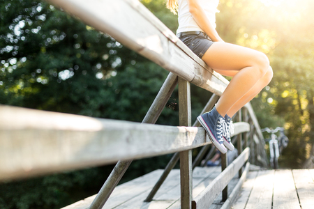 Young woman with beautiful sporty legs sitting on a wooden bridge railing in jeans sneakersの写真素材