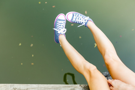 Young woman wearing jeans sneakers sitting in a river bank hanging her  legs over the water