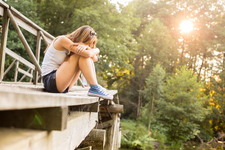 Young sad woman with beautiful sporty legs sitting on a wooden bridge railing in jeans sneakersの写真素材