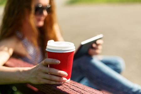 Young woman with red disposable paper cup of coffee using tablet pc sitting on a red bench in a park の写真素材